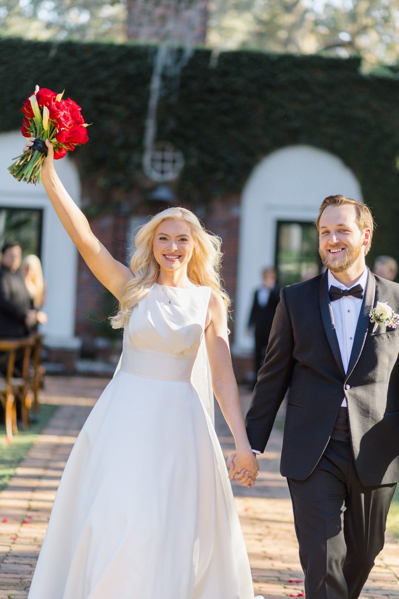 Bride raises her red bouquet as she and the groom walk back up the aisle hand in hand