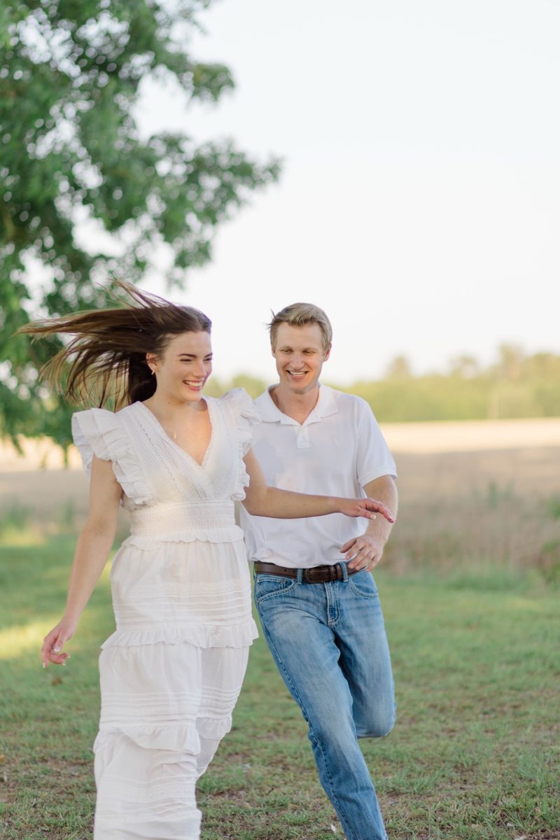 Fiancée runs across a lawn with her hair flying, her fiancé laughing beside her