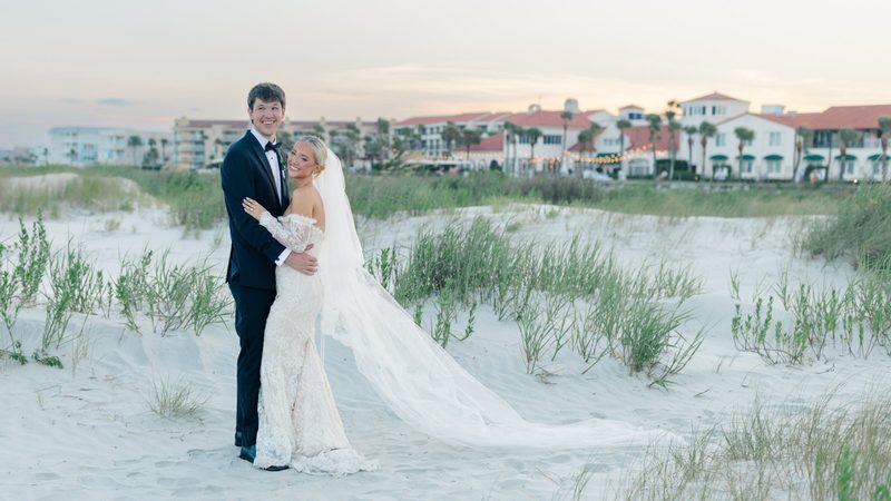 Bride and groom embrace on the sand dunes at sunset, her long veil trailing behind them