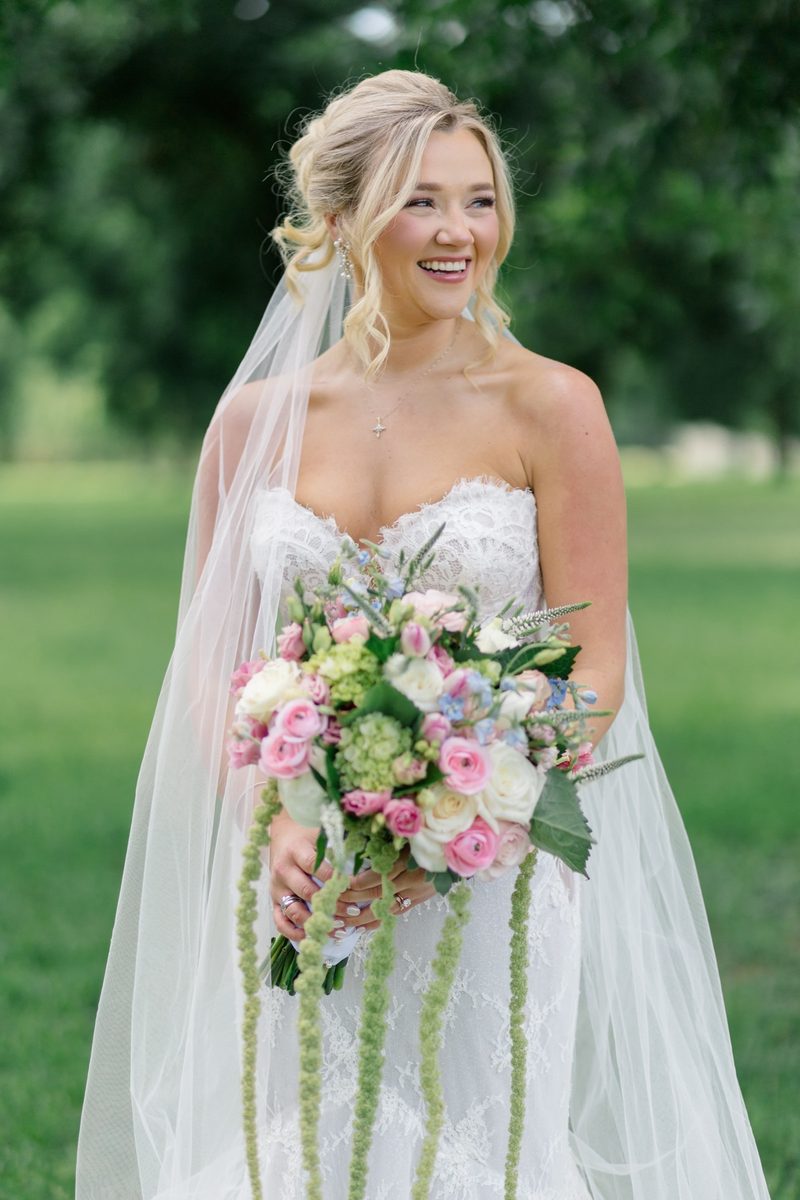 Bride smiles over her shoulder, holding a pink and green bouquet, her veil falling past her gown