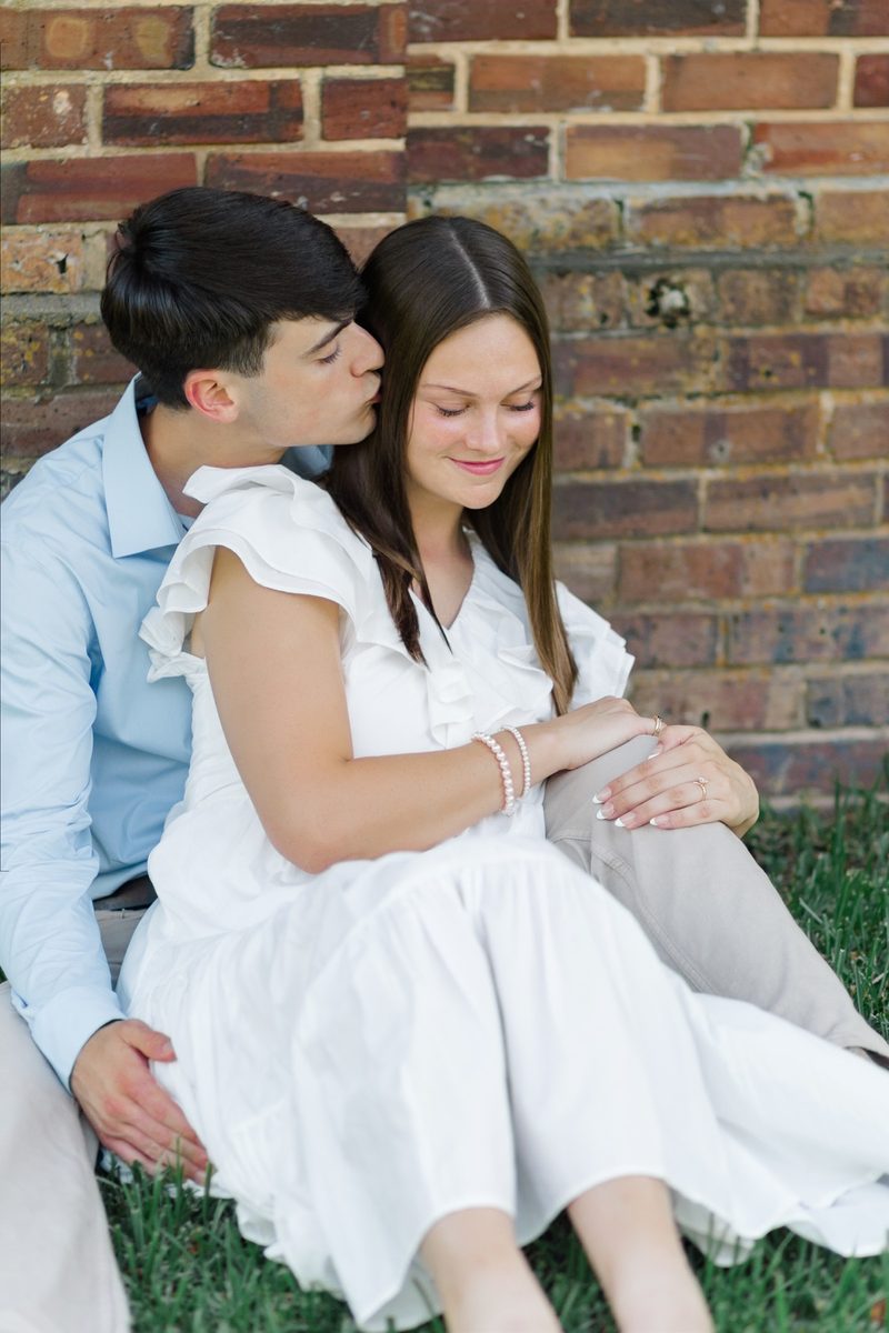 Fiancé kisses her cheek as they sit against an old brick wall