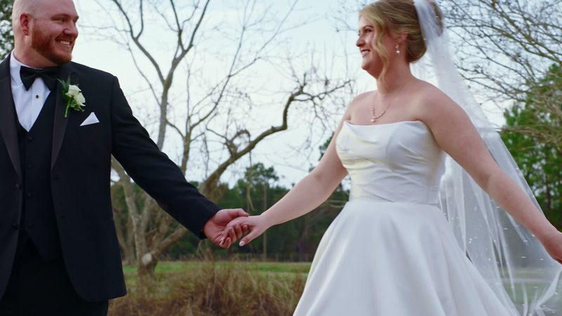Film still: groom leads his bride by the hand across a field, her veil lifting in the breeze
