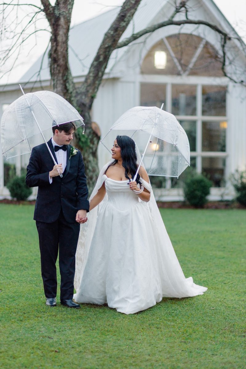 Couple walk across a lawn under clear umbrellas toward a white chapel in the rain
