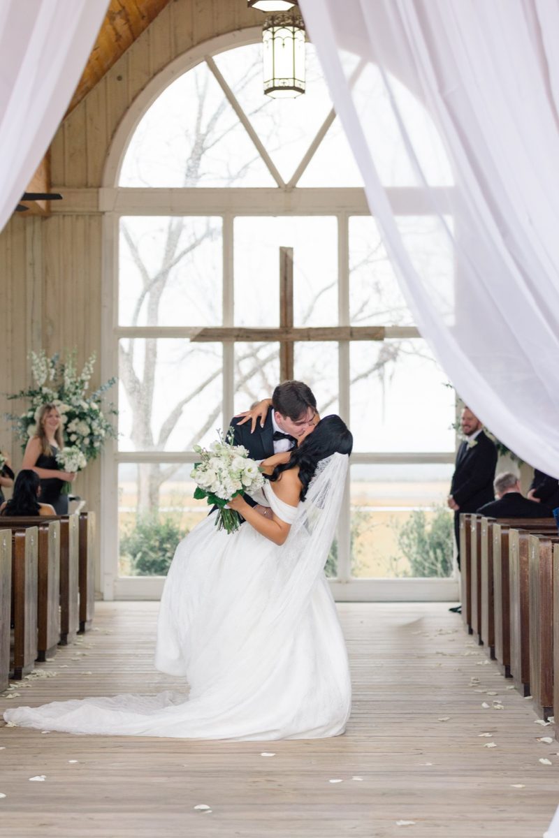Couple kiss at the end of the aisle in a chapel with a cross in the window