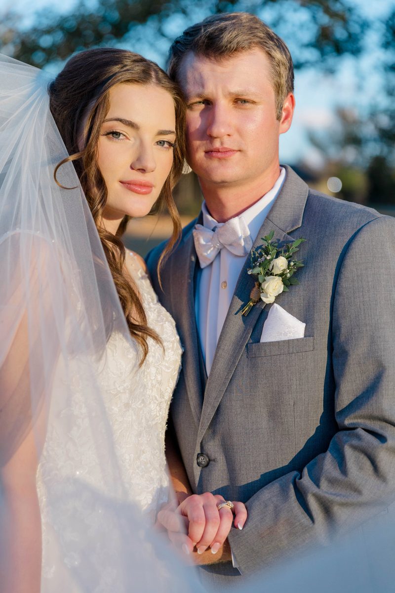 Bride and groom pose close together at golden hour, her veil drifting across her shoulder