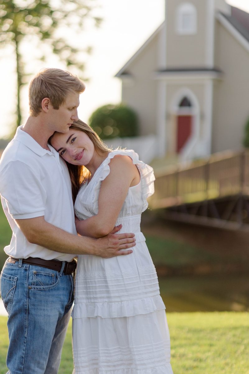 Fiancé rests his chin on her head as she leans into him beside a small chapel