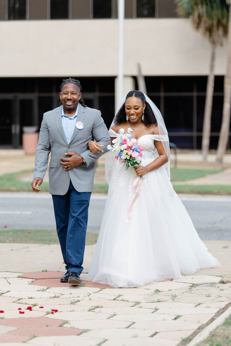 Bride in an off-the-shoulder gown walks down the aisle on her father's arm, bouquet in hand