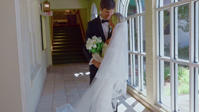 Film still: groom and bride in a sunlit hallway, her veil and bouquet beside tall windows
