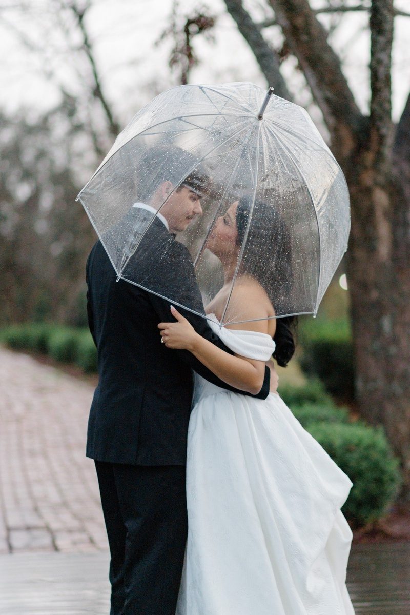 Couple embrace under a clear umbrella on a rainy wedding day