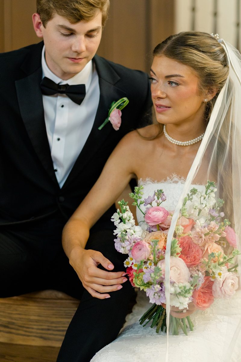 Bride and groom in a black tux sit close together on a church pew, her bouquet in her lap