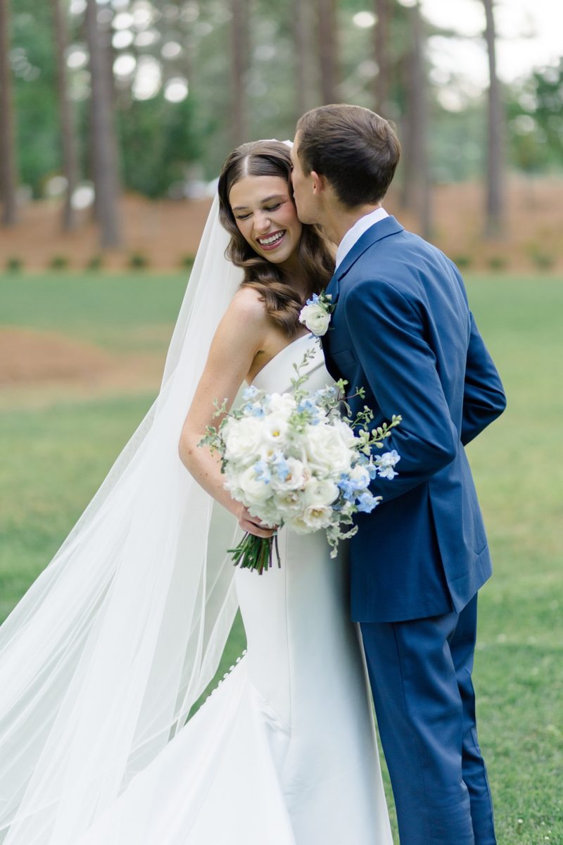 Groom kisses the bride's cheek as she laughs, her long veil trailing across the lawn