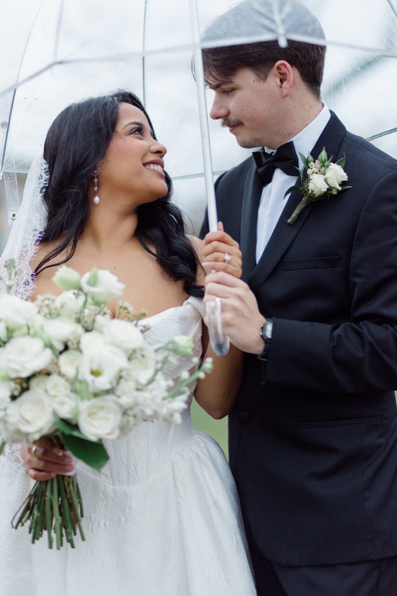 Couple smile at each other under a clear umbrella, she holds white ranunculus