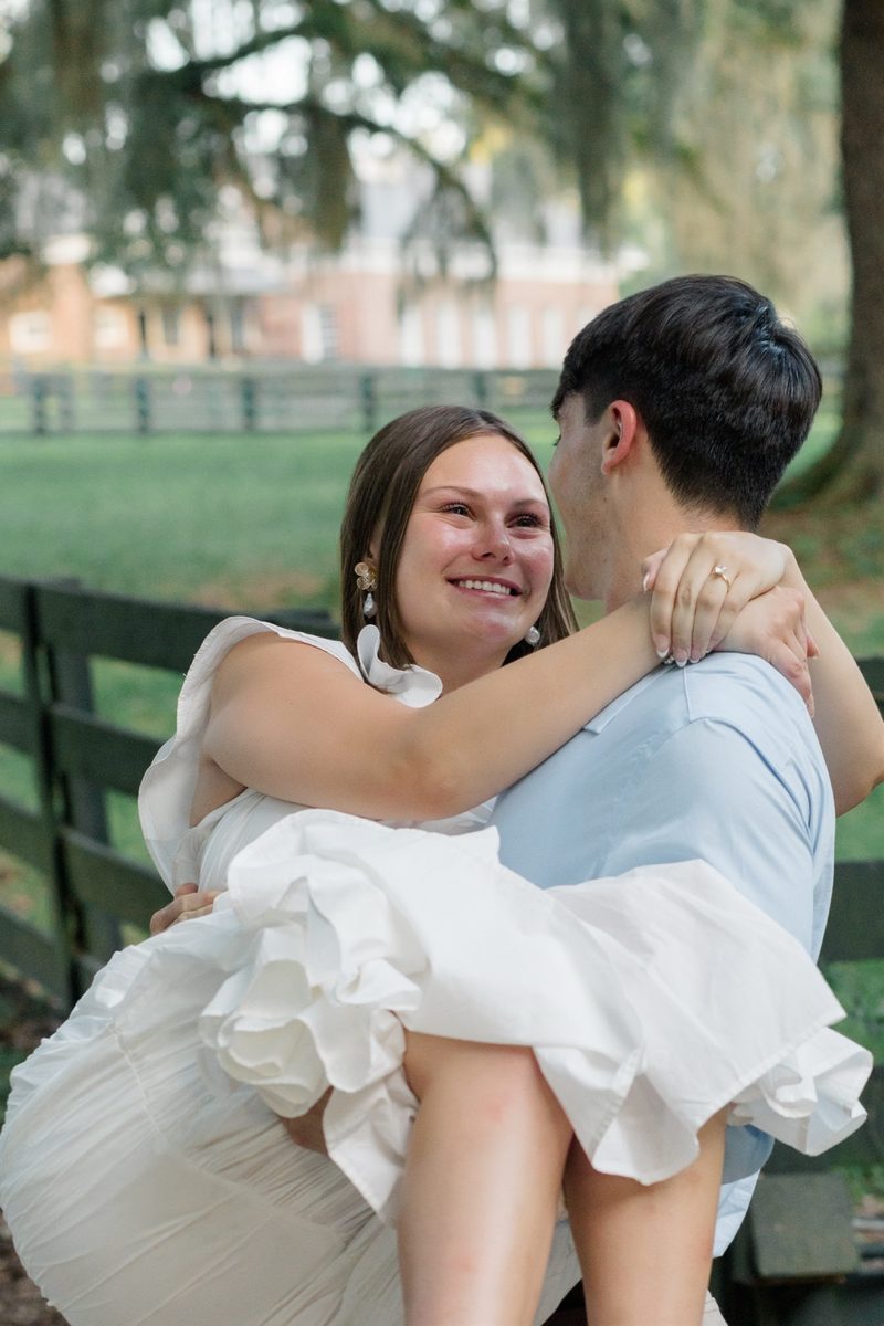 Fiancé lifts her in his arms beside a wooden pasture fence, both laughing