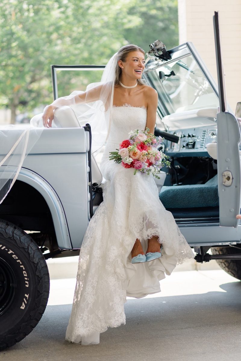 Bride in a lace gown steps out of a vintage blue Bronco, bouquet in hand