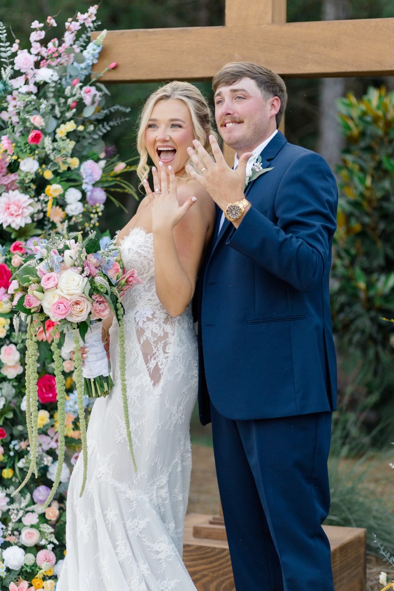 Bride and groom hold up their rings in front of a wildflower-covered ceremony cross