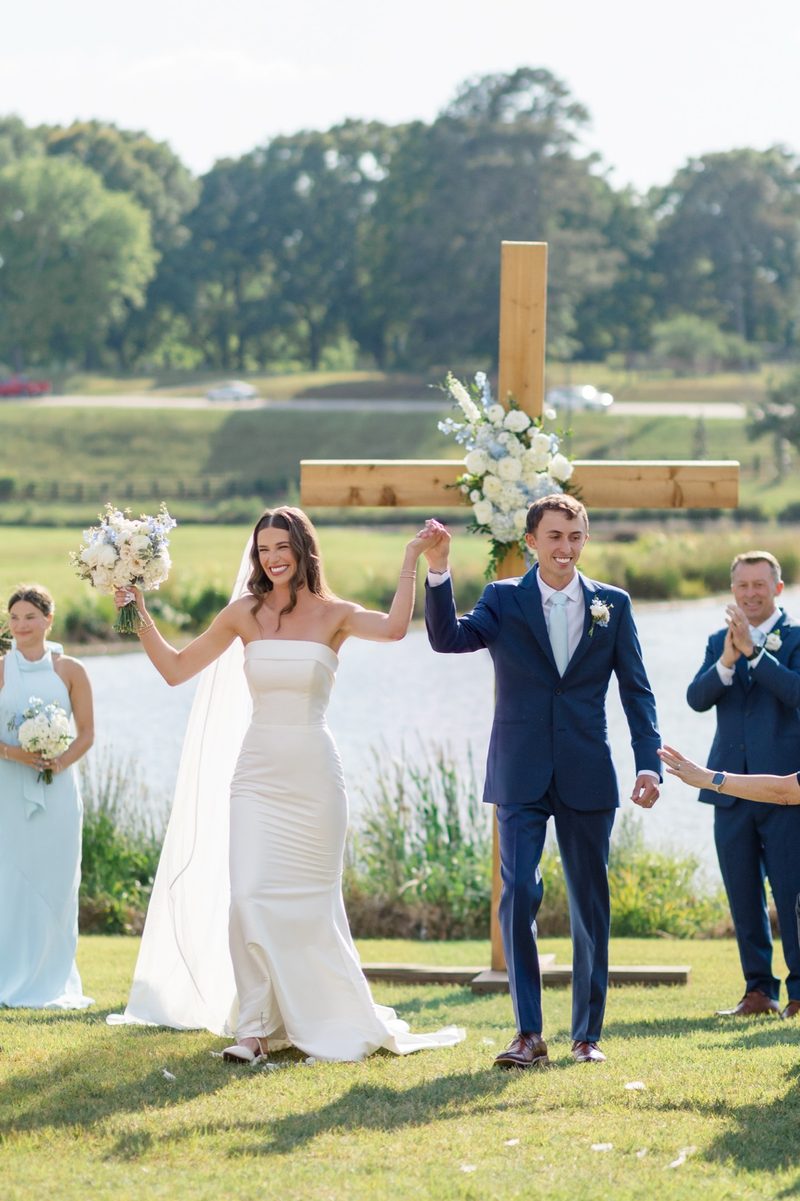 Couple raise their joined hands after the ceremony beside a lake and wooden cross