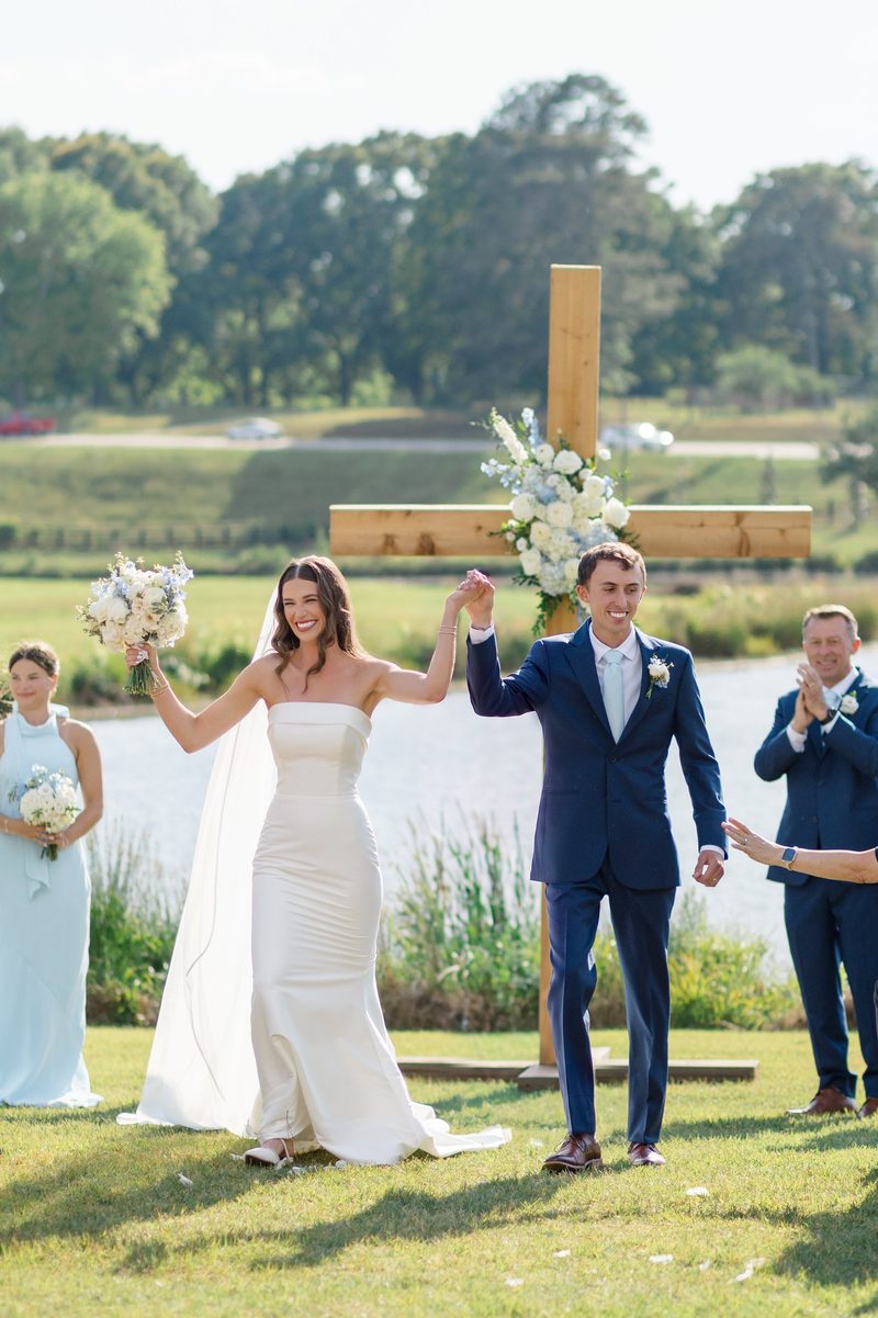 Couple raise their joined hands at the end of the ceremony beside the pond and wooden cross